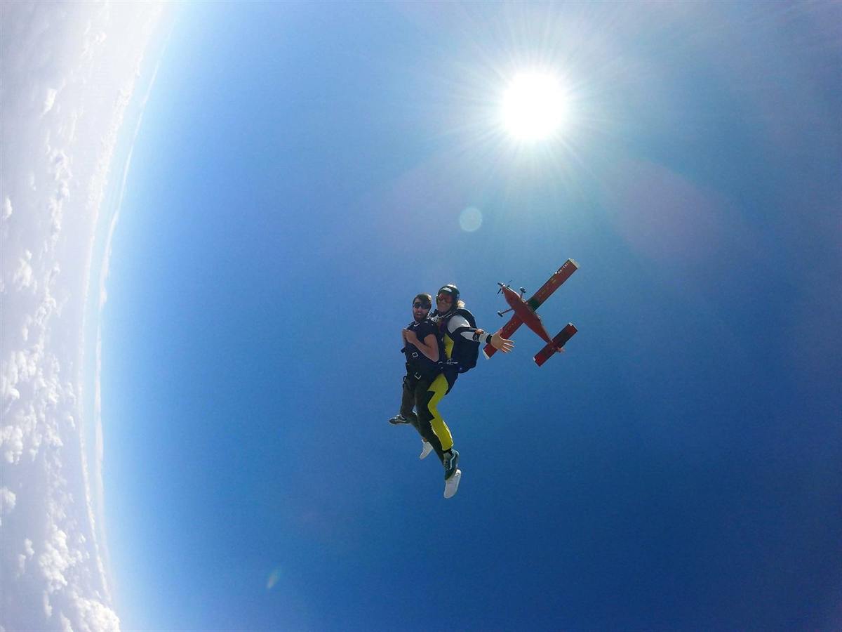 Pareja en salto tándem en caída libre, con el avión al fondo